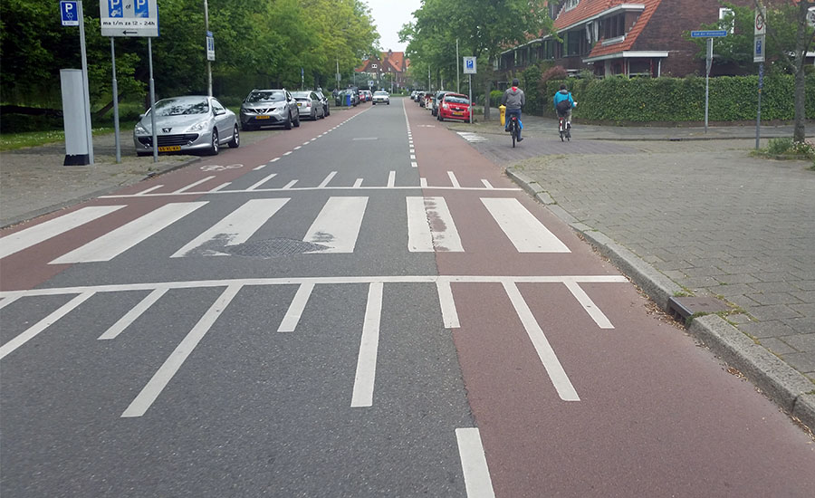 A Delft-area street with on-road parking, dedicated red bicycle lanes, and a single lane for vehicle traffic. Pedestrian crosswalks are raised to help keep traffic moving fairly slow.