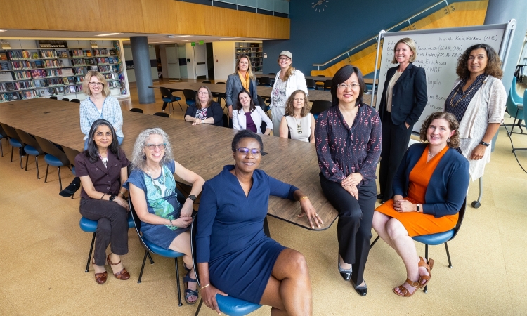 13 women in professional attire pose around a table in an academic space