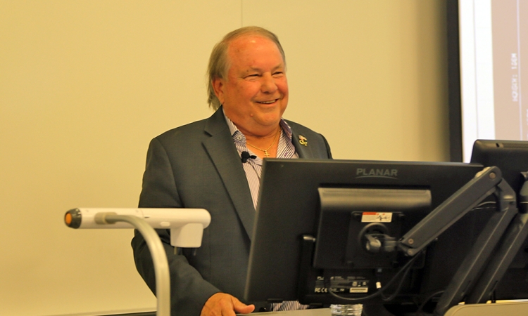 John Carroll in a navy blue blazer and a lavender dress shirt standing at the podium delivering a lecture