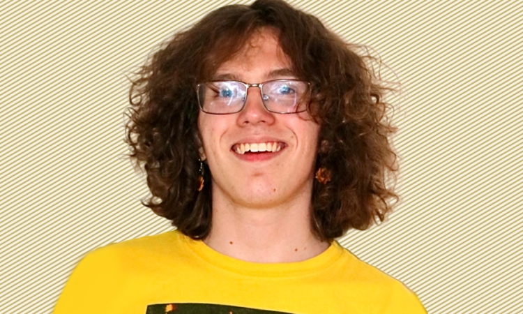 A photo of Hudson McGaughey wearing a yellow T-shirt and glasses in front of a gold and white striped background