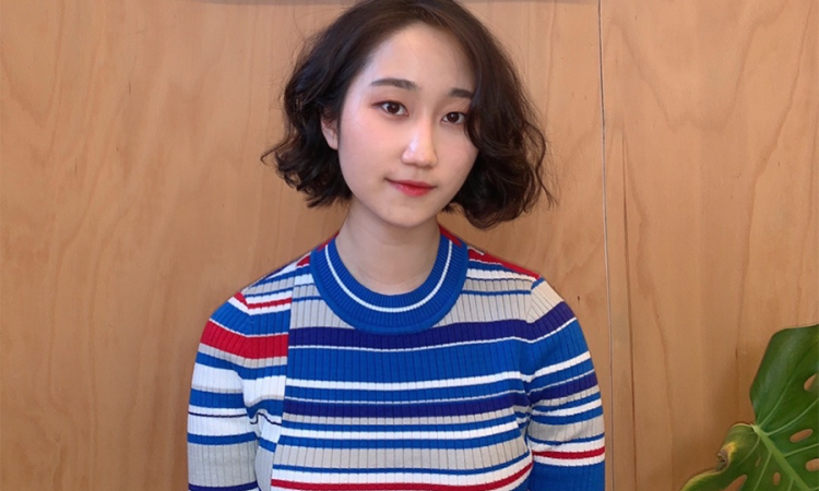 Laura Yang wears a striped sweater and poses in front of a wooden background with a plant visible in the lower right corner