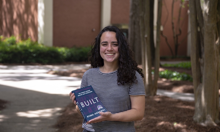 Maria Warren holds the book "BUILT" by Roma Agrawal standing in front of trees and a brick building.