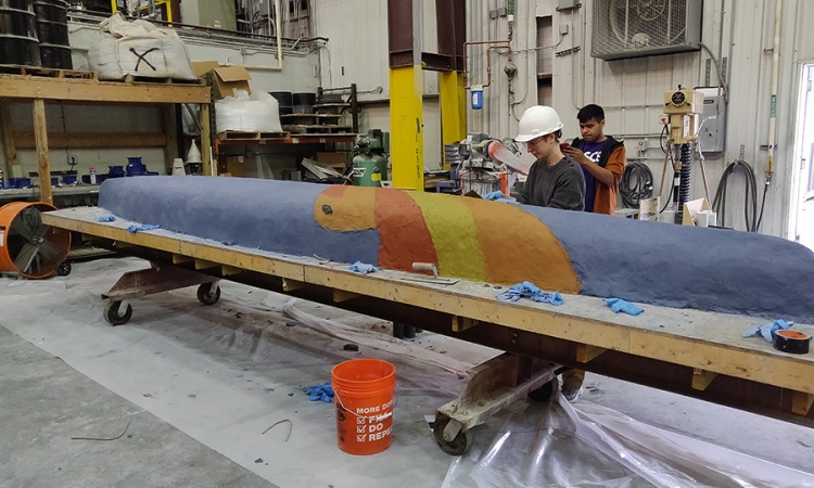 A person in a white hard hat works on a blue canoe with red orange and yellow stripes 
