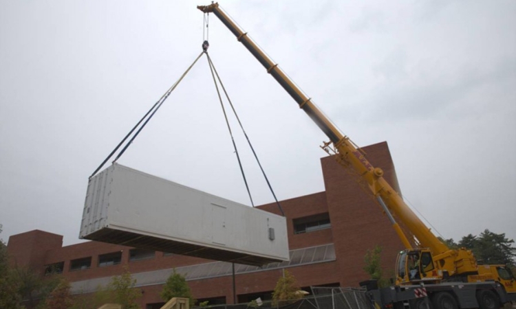 A yellow lift drops a white shipping container next to a brick building. 