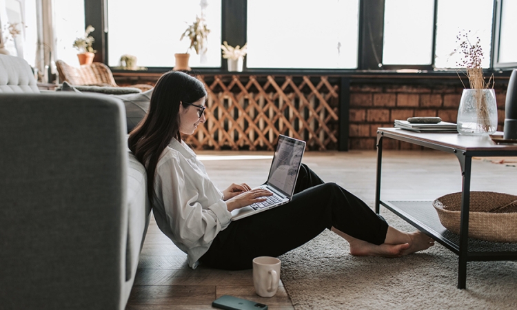 A woman sitting with a laptop computer on her lap and a cup of coffee on the floor of her home