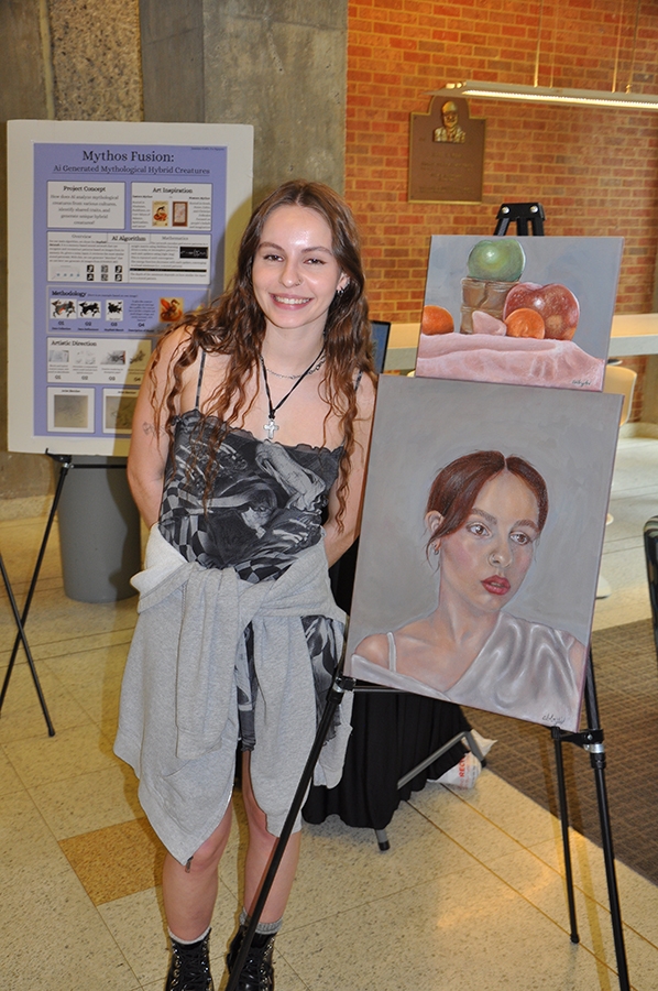 Female student in a dress standing next to an easel with a still life at top and a self-portrait