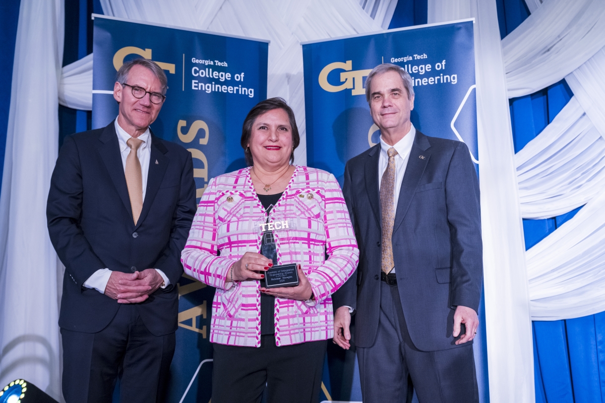 A woman in a pink jacket holds an award between two men in suits