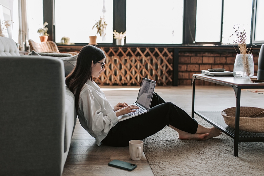 A woman sitting with a laptop computer on her lap and a cup of coffee on the floor of her home